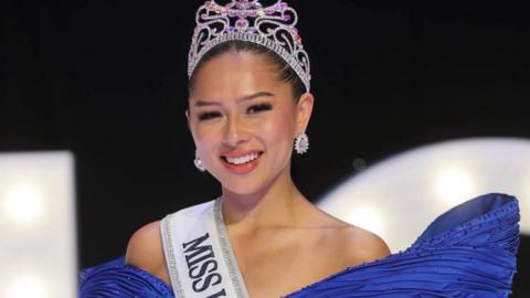 Angela Aumonier has dark hair tied back, is wearing a blue ball gown, a white sash over the shoulder where you can see the word 'miss' in black and is wearing a jewelled tiara.