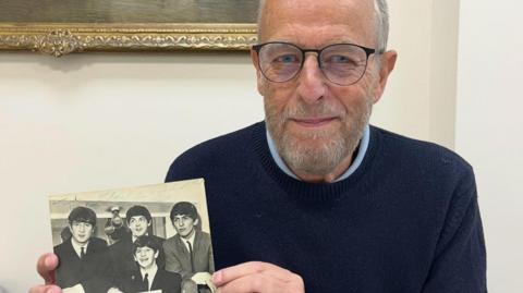 A man in a navy jumper, black glasses and a light blue shirt is holding a black and white signed photo of the Beatles 