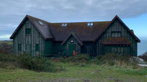 A green wooden building on a cliff face, with moss growing on the roof. 