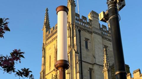 A large white cylinder light is off-centre to the lef in the foreground. Next to it is a black pole. In the background are ornate university buildings.