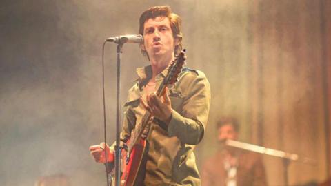 A man with brown wavy hair on stage, standing behind a microphone and holding a guitar. He is looking into the crowd.