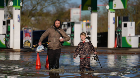 A man and a young boy carrying fishing rods wade through knee-high floodwater near petrol station pumps 