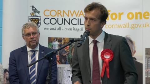 Labour's Noah Law giving a speech in front of a microphone dressed in a grey suit with Steve Double behind him to the left in a blue suit and tie watching on. 