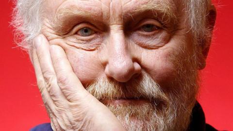 A close up shot of Hodges. He has white hair, a white/brown beard, and blue eyes. He has a hand on his cheek.