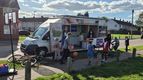 A group of children playing with their bikes and footballs close to a van parked on a residential street. There is basketball hoop close by. The van doors are opened with two people standing inside