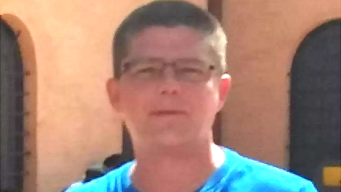 A man with close cropped hair, glasses and a blue shirt. He is looking straight into the camera while standing outside.