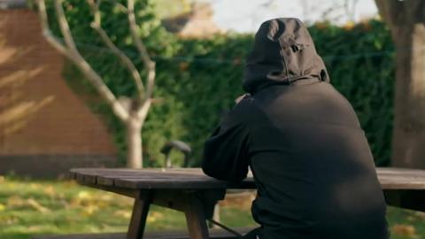A man the BBC is calling Hakan sitting at a picnic bench, pictured from behind so you cannot see his face. He is wearing a black coat with the hood up.