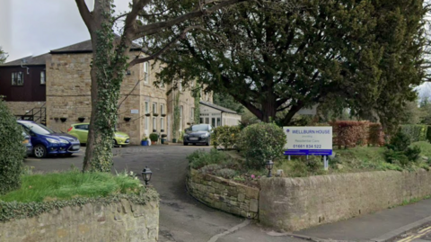 Wellburn House. A large two-storey building made from beige stone with an extension to the rear, the upper floor of which is clad in dark wood. A wide driveway leads to it from the road, with hedges and trees in front. A white and blue sign identifies the facility.