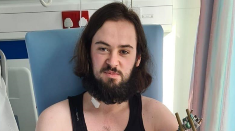 A young man, with long dark brown hair and a brown beard and moustache , sits next to a hospital bed. He has a bandage on his neck.