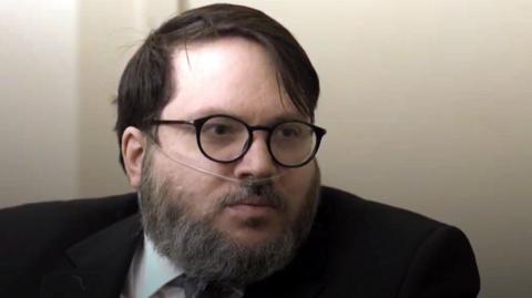 A bearded man with an oxygen tube in his nose. He is wearing glasses and a dark suit