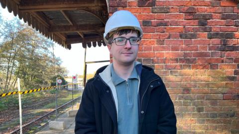 Alex Bramer from The Middleton Towers Restoration Group stands in front of a red brick wall. He is wearing a white hard hat, dark blue coat and light blue fleece underneath. 