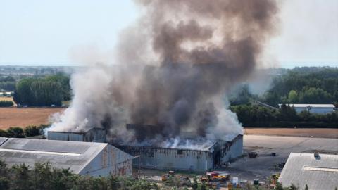 A metal warehouse with a plume of smoke coming from the roof. Fire engines can be seen parked next to the building.