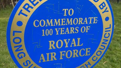 A blue circular sign with gold writing on. Its a plaque commemorating 100 years of the Royal Air Force. Nazi Swastikas have been engraved onto it. 