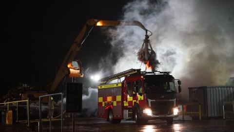 A fire engine in front of a fire. There is lots of smoke against a night sky. A crane is next to the fire engine lifting flaming materials into the air.