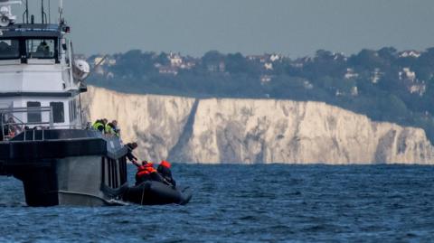 A small boat containing migrants in the English Channel off the coast of Dover receiving support from a larger Border Force boat. The white cliffs are in the background