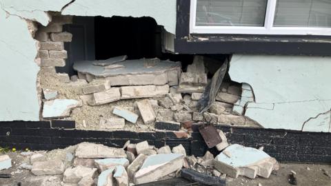 To the left hand side of the a broad living room window, on the external wall of a house, there is a hole in the wall through which you can see the inside of the house. Broken bricks have spilled onto the driveway.