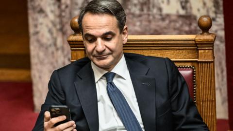 Greek Prime Minister Kyriakos Mitsotakisis wearing a suit and sitting down, looking at a cellphone he holds in his right hand