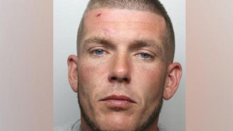 A custody photo shows a man with a shaved head and blue eyes and a cut on his forehead. He is wearing a grey t-shirt.