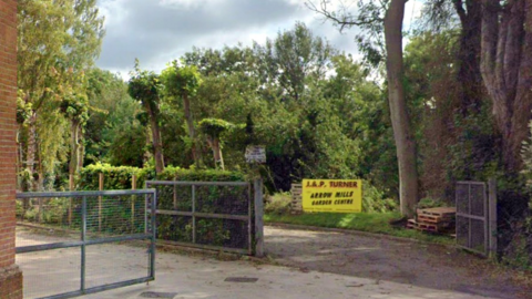 The entrance to the garden centre has a gate which is open in the photo. There is a metal fence on the left with a hedge next to it and another gated entrance further left. A yellow sign is at front of the entrance.