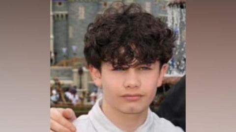 A young boy wearing a white polo shirt. He has curly brown hair. Someone has their hand on his shoulder. Behind, out of focus in the background, is a medieval castle-looking building.