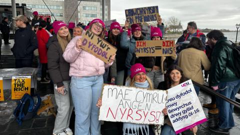 Protesters outside the Senedd in Cardiff Bay wearing pink UCU union-branded hats and holding signs and banners in Welsh saying: no nurses=no NHS and "Wales needs nurses"