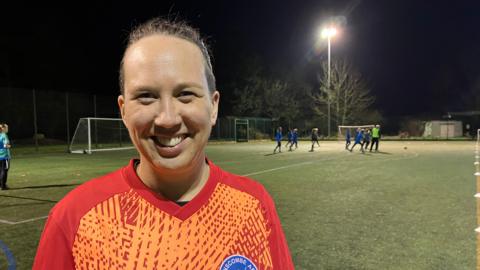 A women is smiling at the camera. She is standing on a football pitch with players in the background. The pitch is lit with floodlights. She has brown hair tied back and is wearing an orange patterned football top. 