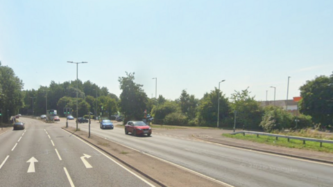 The A602, showing a dual-carriageway with cars on the road and a supermarket to the right. There is a junction with road markings on it. Trees are by all the entrances.