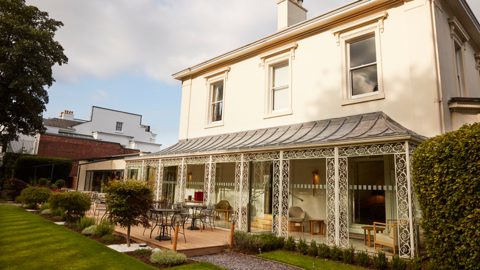 Cream-coloured two‑storey Georgian building with an ornate ironwork veranda overlooking a terrace with seating and lawn. Three sash windows are on the second story and ornamental trees and bushes line the garden.