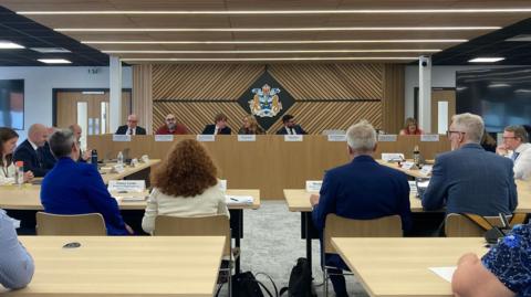 The council chamber at Dunedin House has several wooden tables and chairs facing a wooden panel. Seven people are sitting on the panel, behind white named cards. Behind them, is the blue and gold council crest.