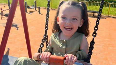 Niamh is photographed on a swing in playpark. She has her hair in two pigtails at the sides, held in place by pink bows. She is smiling. She has blue eyes and is wearing a green top.