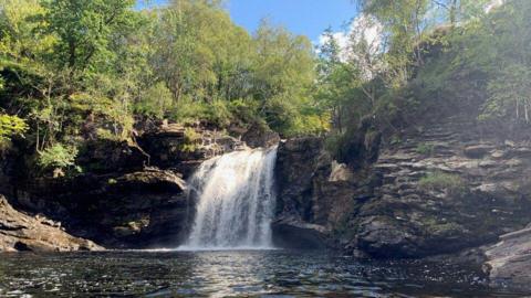 A small waterfall cascades over a rocky cliff into a clear, calm pool surrounded by steep rock walls and dense green trees. Sunlight filters through the foliage, reflecting on the water's surface and illuminating the layered stone formations along the shoreline.