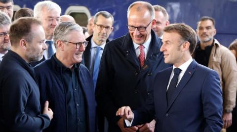 Starmer, Macron and Merz at Kyiv railway station, smiling with people behind them.