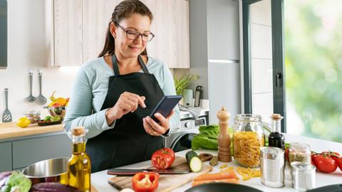 A woman in a kitchen wearing an apron and looking at a mobile phone. In front of her is a kitchen counter covered in vegetables, a bottle of olive oil and a jar of pasta.