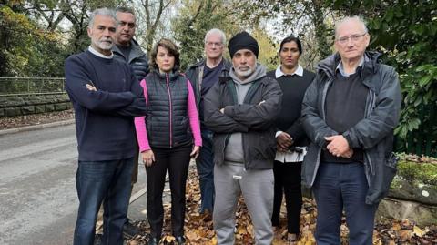 A group of residents standing on a road, with no pavement, looking at the camera. There are trees and autumn leaves on the ground
