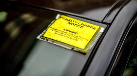 A yellow penalty charge notice stuck to the windscreen of a black car.
