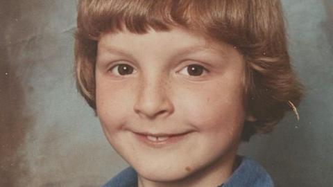 A young boy with light, brown hair, brown eyes and freckles over his nose smiles into the camera. His two front teeth slightly protrude over his bottom lip
