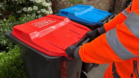 A worker in an orange hi vis jacket approaches two household recycling bins, one red and one blue.