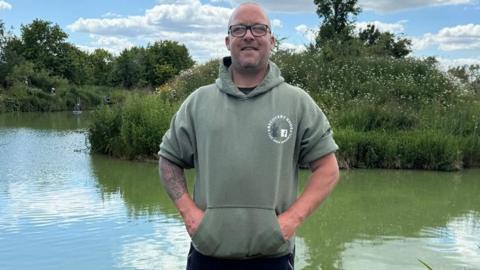 A man wearing glasses and a green hooded top with the sleeves rolled up stands in front of a fishing lake. The top has a circular logo for the group Recovery Rods