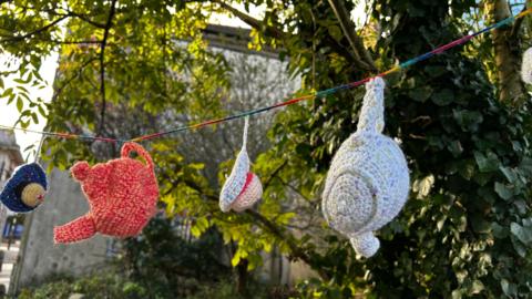 Knitted teapots and mugs tired to rainbow strong. In the background is a tree with green leaves. 