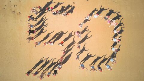 An aerial image looking down onto a sandy beach. Multiple members of the Jersey Long Distance Swimming Club are stood on the sand in the shape of a number 50 with their hands in the air.