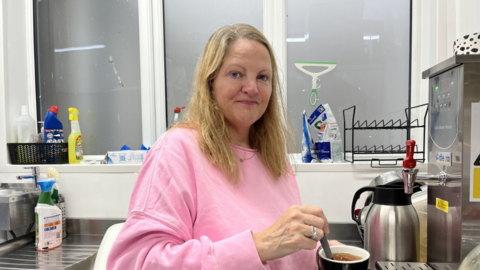A woman in a pink jumper with long blonde hair makes a cup of tea in a kitchen, with sink and cleaning products visible in the background under a window with privacy film on the glass. A hot water tap can be seen in front of her and a rack for dishes on the window ledge.
