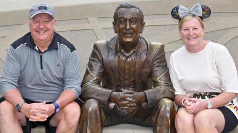 Phil Taylor has a grey cap, grey top and dark shorts and is sitting with Melanie Taylor, who has long blonde hair tied back, white top with pattern at the bottom and black shorts/skirt. They are both sitting down and between them is a bronze statue of Walt Disney.
