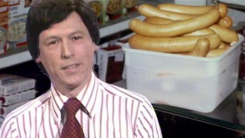 News reader sits in front of an image of Swedish sausages.