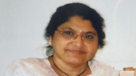 An older looking woman is seen in closeup. She wears earrings, and has dark hair and glasses.
