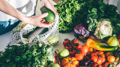 Young woman takes fresh colourful vegetables out of mesh bag putting on kitchen table at home.