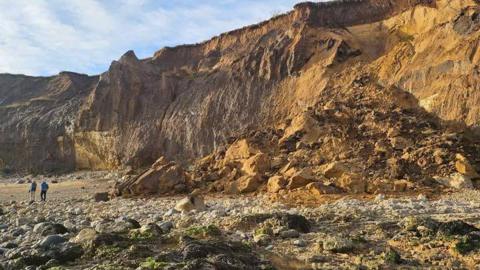 The face of a big, brown cliff with a pile of rocks slumped at the bottom of the right side. Two people can be seen walking along the beach in front of the landslip