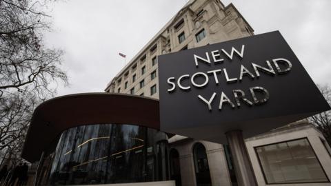 The outside of New Scotland Yard. The name of the station is written in silver letters on a black board outside a large cream building