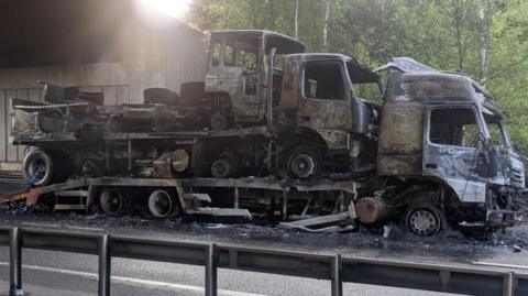 Charred shell of lorry with a further two being carried on its trailer.  The lorry is at the side of a road under a bridge