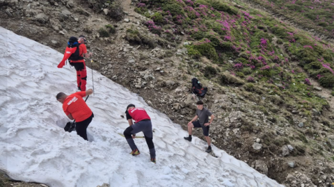 A rocky hillside with people in red coats on an area covered in snow.