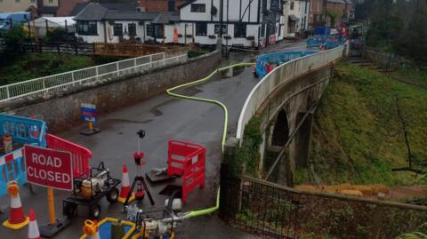A bridge with buildings on the opposite side. There are road closed signs and traffic cones and various pieces of machinery as well as blue temporary fencing on the bridge. There are fixed white rails either side of the bridge and a large grass embankment can be seen on the far side.